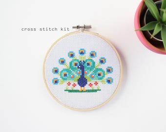 May include: A cross stitch kit featuring a peacock design. The peacock is rendered in blue, teal, and orange cross-stitch on white fabric, set in a wooden hoop. The text "cross stitch kit" is above the hoop.