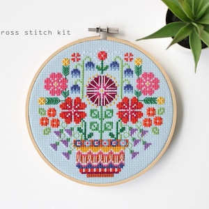May include: A cross stitch kit with a floral design in a wooden hoop. The design features pink, red, and yellow flowers, green leaves, and blue accents on a light blue background. The text "cross stitch kit" is visible.