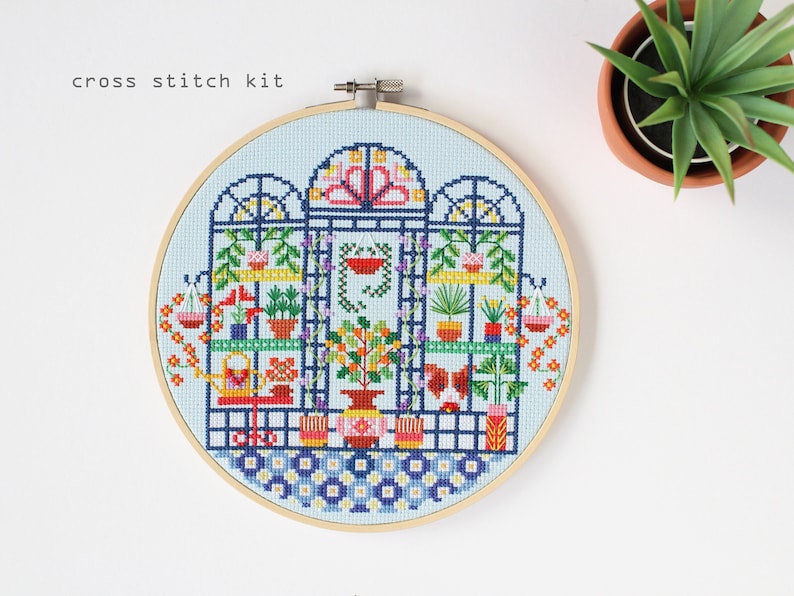 May include: A cross stitch kit featuring a colourful illustration of a greenhouse with plants and flowers. The kit includes a hoop, fabric, and floss.