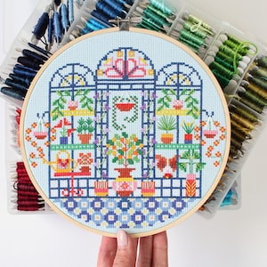 May include: A colorful cross-stitch embroidery hoop depicting a greenhouse with plants and flowers. The hoop is made of wood and has a blue background.