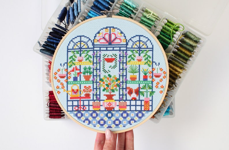 May include: A colorful cross-stitch embroidery hoop depicting a greenhouse with plants and flowers. The hoop is made of wood and has a blue background.