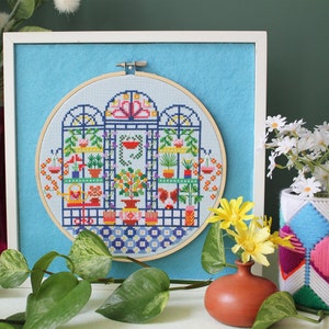 May include: A framed cross-stitch embroidery depicting a colourful indoor garden scene with plants, flowers, and a watering can. The embroidery is in a round hoop and is set against a light blue background.