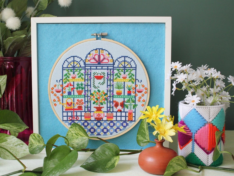 May include: A framed cross-stitch embroidery depicting a colourful indoor garden scene with plants, flowers, and a watering can. The embroidery is in a round hoop and is set against a light blue background.