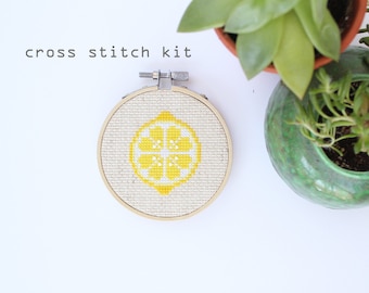 Puede incluir: Un kit de punto de cruz con un diseño de limón amarillo sobre un fondo blanco. El kit está en un aro de madera redondo.