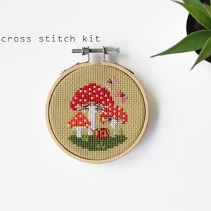 May include: A cross stitch kit featuring a mushroom design. The embroidery hoop displays a red and white mushroom with a green base, and the text "cross stitch kit". The background is a tan fabric.