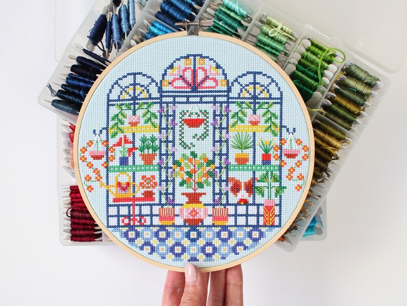 May include: A colourful cross-stitch embroidery hoop depicting a whimsical indoor garden with plants, flowers, and a butterfly. The hoop is surrounded by a variety of colourful embroidery floss spools.