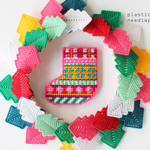 May include: A colourful needlepoint Christmas stocking surrounded by a wreath of plastic canvas Christmas tree shapes in various colours. The stocking is pink, red, yellow, green, and blue. Text reads "plastic canvas needlepoint kit."