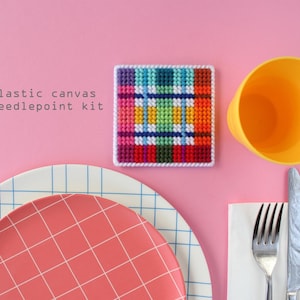 May include: A plastic canvas needlepoint kit with a colorful plaid design. The kit is on a pink surface with a yellow cup, a white napkin, a fork, and a knife.
