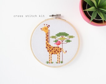 May include: A cross stitch kit featuring a giraffe with a tree and bird design. The giraffe has a yellow body with brown spots, and the tree has green leaves. The text "cross stitch kit" is above the design.