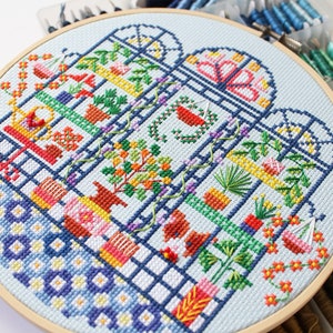 May include: A colourful cross-stitch embroidery hoop depicting a stylised greenhouse with various plants and flowers. The hoop is made of wood and has a light blue background.