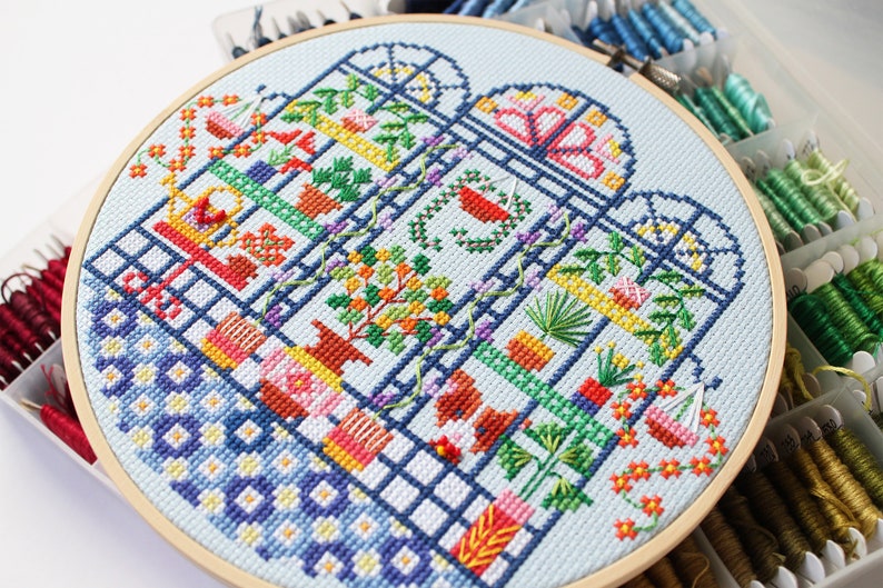 May include: A colourful cross-stitch embroidery hoop depicting a stylised greenhouse with various plants and flowers. The hoop is made of wood and has a light blue background.
