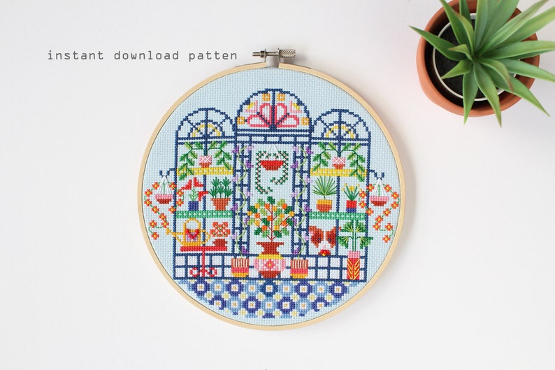 May include: A cross-stitch pattern of a colorful greenhouse with plants, flowers, and gardening tools. The pattern is framed in a wooden embroidery hoop.
