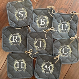 May include: Set of eight gray quilted pot holders with white embroidered letters and green leaf wreaths. The letters are S, B, U, R, J, C, H, and M.