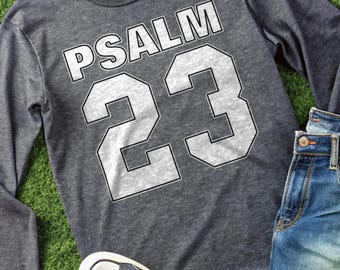 Może przedstawiać: Szara koszulka z długim rękawem z napisem "PSALM 23" w białych literach blokowych. Numer "23" jest wyświetlany pod słowem "PSALM". Koszulka jest połączona z niebieskimi dżinsami i niebieskimi trampkami.