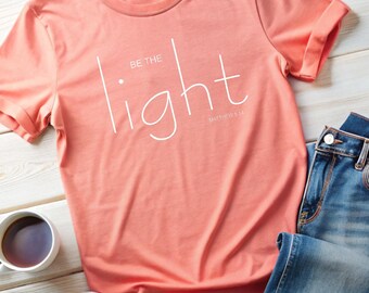 May include: A coral colored t-shirt with the text "Be the light" in white, with the scripture reference "Matthew 5:14" underneath.