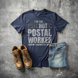 以下が含まれることがあります： 「I'M THE PSYCHOTIC POSTAL WORKER」と「EVERYONE WARNED YOU AP」の文字が入ったネイビーブルーのTシャツ。画像には、茶色の革ブーツと折りたたまれたブルージーンズも写っています。