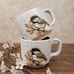 May include: Two speckled white ceramic mugs stacked vertically, each featuring a hand-painted illustration of a black, white, and yellow chickadee perched on a branch with small pink flowers. The mugs have a sturdy handle and a smooth base.