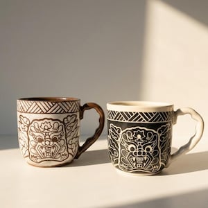 May include: Two ceramic mugs with detailed designs. One is off-white with brown detailing and a brown handle. The other is black with white detailing and a white handle. Both mugs feature a detailed face design and geometric patterns.