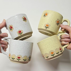 May include: Four ceramic mugs with floral designs. Two mugs are white with red and yellow flowers, and two are yellow with orange and red flowers. Each mug has a curved handle and a speckled finish. The mugs are held up against a white background.
