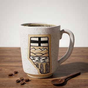 May include: A speckled white ceramic mug filled with coffee, featuring a black and tan graphic of a landscape and a cross. A small wooden spoon and coffee beans are on the wooden surface.