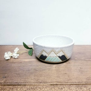 May include: A white ceramic bowl with a speckled glaze features a hand-painted design of three mountains in black, white, and light blue.