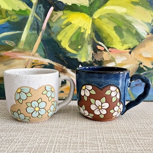 Può includere: Due tazze in ceramica con disegni floreali. Una tazza è bianca con fiori blu su una forma marrone. L'altra è blu scuro con fiori bianchi su una forma marrone. Entrambe le tazze hanno manici.