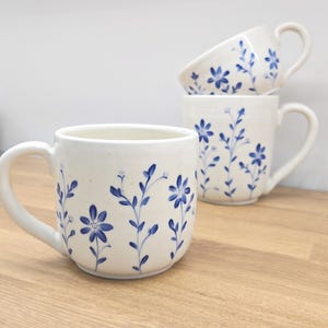 May include: Three white ceramic mugs with blue floral patterns. The mugs have a rounded shape and a handle. The floral design features blue flowers and leaves. The mugs are displayed on a wooden surface.