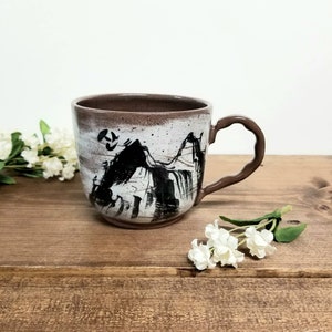 May include: A ceramic mug with a brown handle and rim, featuring a black and white mountain landscape design. The mug is set on a wooden surface, with white flowers in the background. The mug's design is reminiscent of a brush painting.