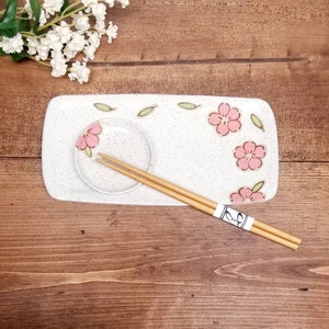 Puede incluir: Bandeja de servir de cerámica blanca con un pequeño cuenco para salsas y un diseño floral con flores rosas y hojas verdes. Dos palillos de madera están colocados en la bandeja.