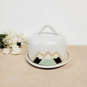 May include: White ceramic cheese dome with a handle and a mountain design in black, white, and green. The dome sits on a matching plate.