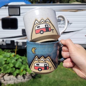Op de afbeelding: Twee keramische mokken met een kampeerthema. De bovenste mok is wit met bruine spikkels en heeft een rode en witte camper voor een bergketen. De onderste mok is blauw met een zwart-witte maan en een rode en witte camper voor een bergketen.