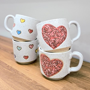 Peut inclure: Quatre tasses en céramique blanche mouchetée empilées sur une surface en bois. Deux tasses présentent un motif de cœur fait de roses roses, tandis que les deux autres ont des contours de cœur colorés. Chaque tasse a une anse incurvée.