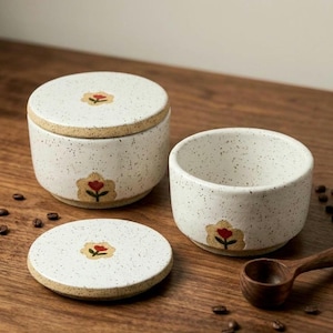 May include: A set of speckled ceramic kitchenware, including a lidded jar, a small bowl, and a coaster. Each piece features a hand-painted red flower design. The items are on a wooden surface with coffee beans and a wooden spoon.