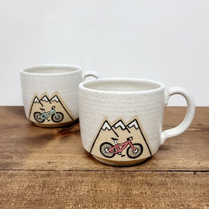 Könnte beinhalten: Zwei weiße Keramikbecher mit gesprenkeltem Überzug. Jeder Becher zeigt ein Bergpanorama mit einem Fahrrad davor. Ein Fahrrad ist türkis und das andere ist rosa.