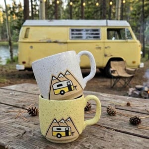 Può includere: Due tazze di ceramica maculata impilate su un tavolo di legno, ognuna con un disegno di montagna e camper. Le tazze sono bianche e gialle, con una grafica di camper gialla. Un camper giallo è sullo sfondo.
