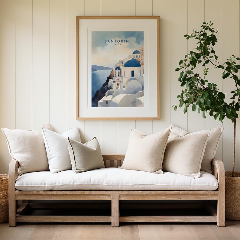 Puede incluir: Una impresi&oacute;n en acuarela enmarcada de Santorini, Grecia, con edificios blancos y c&uacute;pulas azules. Cuelga sobre un banco de madera con cojines y almohadas beige. Una planta en maceta est&aacute; a la derecha. El texto "SANTORINI GREECE" es visible.