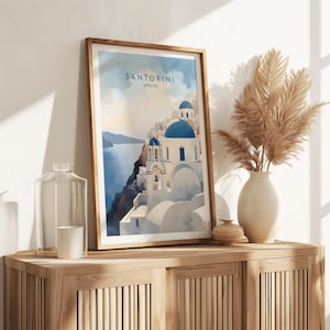 Puede incluir: Una impresi&oacute;n art&iacute;stica enmarcada que representa Santorini, Grecia, con edificios blancos, c&uacute;pulas azules y el texto "SANTORINI GREECE". La impresi&oacute;n se exhibe en un mueble de madera, junto a un jarr&oacute;n con hierba de la pampa seca.