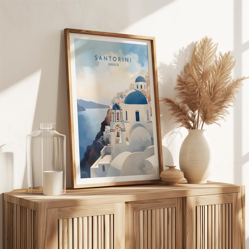 Puede incluir: Una impresi&oacute;n art&iacute;stica enmarcada que representa Santorini, Grecia, con edificios blancos, c&uacute;pulas azules y el texto "SANTORINI GREECE". La impresi&oacute;n se exhibe en un mueble de madera, junto a un jarr&oacute;n con hierba de la pampa seca.