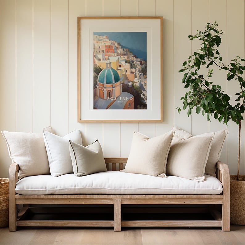 May include: A white linen sofa with a wooden frame and four pillows. A framed print of a colorful cityscape with the text "Positano, Italia" hangs on the wall behind the sofa. A potted plant with green leaves is on the right side of the image.