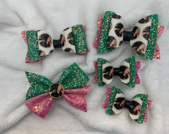 May include: Five glitter hair bows in various sizes. Each bow features a green glitter top layer, a pink glitter bottom layer, and a center image of a woman in a black and white polka dot pattern. The bows are on a white, fluffy surface.