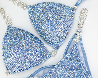 May include: A light blue competition bikini with crystal embellishments. The bikini top has a halter style strap and the bottom has a high cut leg.
