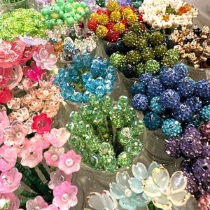 May include: A vibrant display of colorful beaded floral arrangements in various hues, including pink, green, blue, and yellow. The flowers are arranged in small glass containers, creating a visually appealing and decorative display.