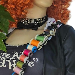 May include: A black top hat with a 10/6 card, orange curly hair, and a black choker. A black shirt features the text "We're All Mad Here" and a graphic of a wide grin. A bandolier of colorful thread spools is worn across the chest over a black jacket.