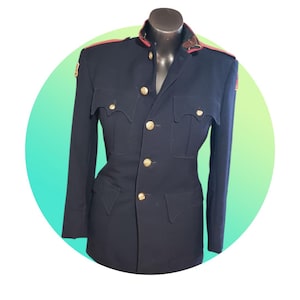May include: A dark navy blue military-style jacket featuring a high collar with red trim, gold-toned buttons down the front, and four scalloped-flap pockets on the chest and waist. The shoulders are accented with red epaulets and small patches.