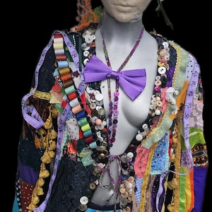 May include: A mannequin wearing a multi-coloured patchwork jacket adorned with numerous buttons, spools of thread, and a purple bow tie. A purple hat sits on the head, and a measuring tape is draped over the shoulders and jacket front.