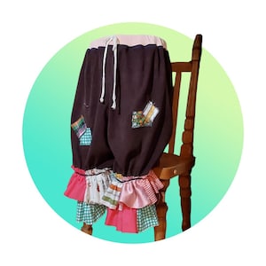 May include: Dark brown pants with colorful patchwork and ruffled cuffs. The pants have a drawstring waist and are decorated with patches of various patterns and colors. The cuffs feature layers of pink, white, and green fabric. The pants are displayed on a wooden chair.