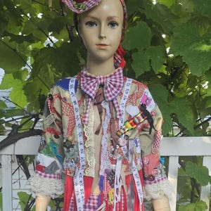 May include: A mannequin wearing a red and white gingham top hat, a matching bow tie, and cropped trousers. The jacket features a patchwork design with lace trim, various buttons, ribbons, and a white measuring tape draped over the front.