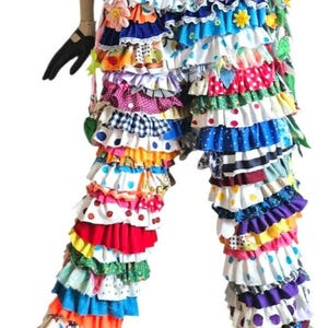 May include: Multi-colored pants featuring numerous horizontal fabric ruffles in various patterns including polka dots, stripes, and floral prints. The garment includes decorative fabric flower appliqués and a yellow elasticized waistband.