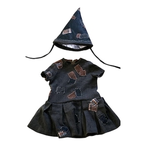 Peut inclure: Une tenue de poupée comprenant une robe gris foncé avec une jupe plissée et un chapeau pointu assorti. La robe et le chapeau sont ornés de divers patchs bruns et bleus, créant un motif patchwork.