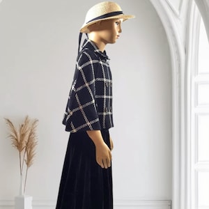 May include: A mannequin wearing a straw hat with a black ribbon, a black and white plaid cape with a collar and buttons, and a long black skirt. The cape features a prominent grid pattern, while the skirt is solid black and reaches the floor.